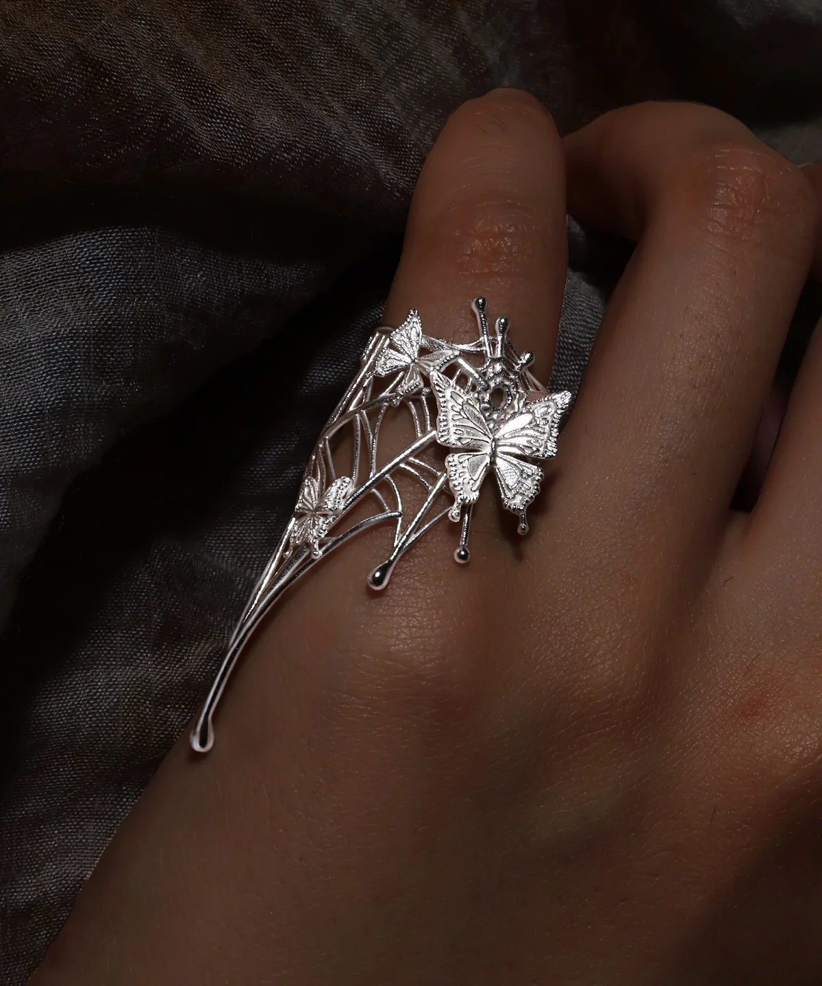 Butterfly Ring in 925 Sterling Silver | Art Fantasy Pinky Ring features an intricately designed silver band adorned with delicate butterfly motifs and a subtle web-inspired pattern. Captured in a close-up on a model’s hand against a dark, textured backdrop, the ring’s shimmering details stand out beautifully. This whimsical yet elegant piece exudes refined craftsmanship, making it a perfect statement accessory for both casual and formal wear, ideal for fashion-forward trendsetters.