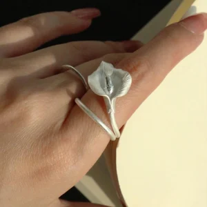 A close-up of a hand wearing the Calla Lily Ring while holding an open book. The handcrafted 999 silver ring features a lifelike calla lily flower that wraps gently around the finger, capturing the soft shimmer of natural light. The elegant floral design stands out against the skin and the pages, highlighting the ring’s delicate texture and sculptural form.