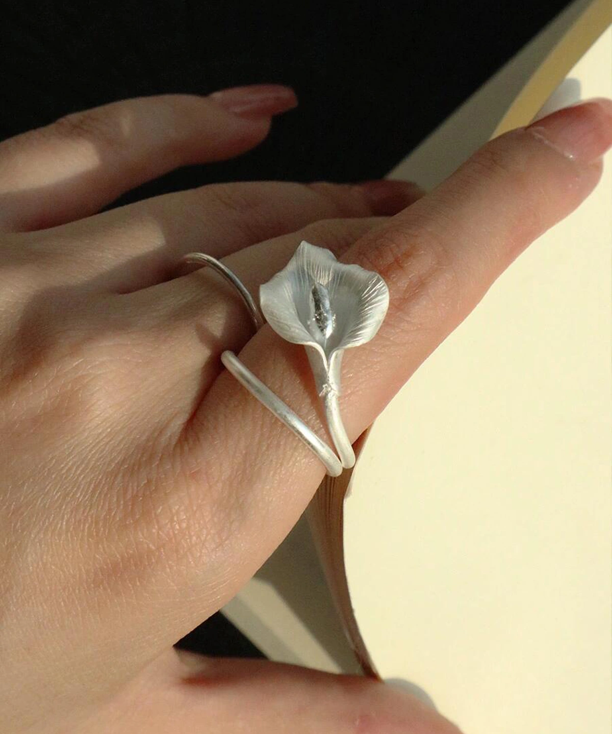 A close-up of a hand wearing the Calla Lily Ring while holding an open book. The handcrafted 999 silver ring features a lifelike calla lily flower that wraps gently around the finger, capturing the soft shimmer of natural light. The elegant floral design stands out against the skin and the pages, highlighting the ring’s delicate texture and sculptural form.