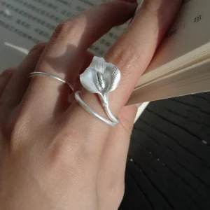 A close-up of a hand wearing the Calla Lily Ring while holding an open book. The handcrafted 999 silver ring features a lifelike calla lily flower that wraps gently around the finger, capturing the soft shimmer of natural light. The elegant floral design stands out against the skin and the pages, highlighting the ring’s delicate texture and sculptural form.