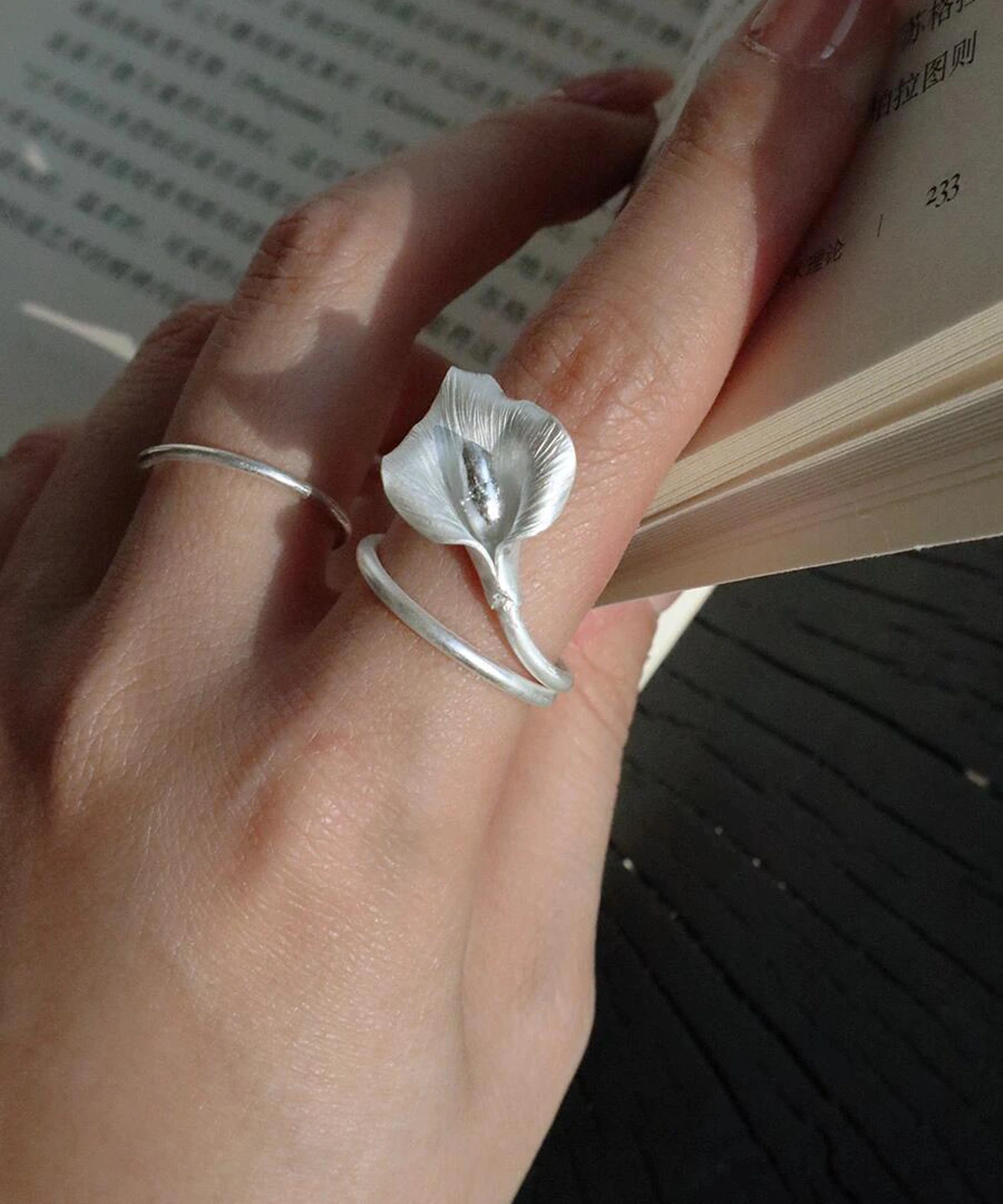 A close-up of a hand wearing the Calla Lily Ring while holding an open book. The handcrafted 999 silver ring features a lifelike calla lily flower that wraps gently around the finger, capturing the soft shimmer of natural light. The elegant floral design stands out against the skin and the pages, highlighting the ring’s delicate texture and sculptural form.