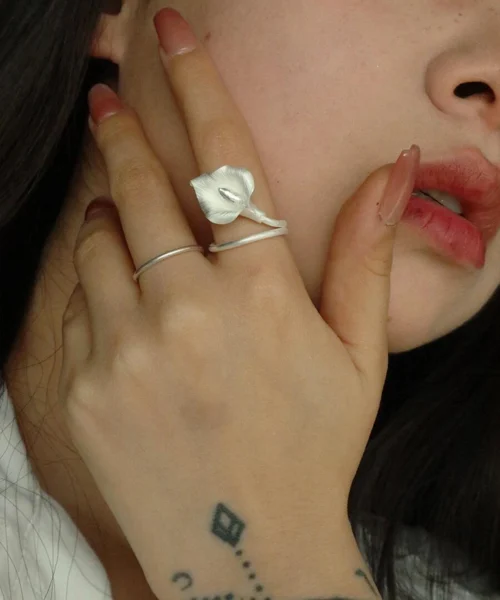 Close-up of a woman wearing the Calla Lily Ring on her index finger. The handcrafted 999 silver ring features a delicate, sculpted calla lily flower with a matte finish, wrapping elegantly around her finger. She gently touches her face with her hand, showing off the ring’s natural curves and detailed petal texture. Her minimalist style and soft expression highlight the organic elegance of the jewelry.