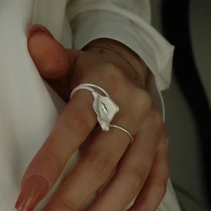 A close-up photograph showcases a hand wearing the Calla Lily Ring, a handmade piece crafted from 999 silver. The ring features an elegant design inspired by the calla lily flower, with a gracefully curved structure that resembles a blooming petal. The silver surface has a refined texture, enhancing its organic and artistic appearance. The adjustable band ensures a comfortable fit for various finger sizes. The hand is set against a soft white fabric background, complementing the delicate and sophisticated aesthetic of the jewelry. This image captures the timeless elegance of the Calla Lily Ring, making it a stunning accessory for jewelry lovers