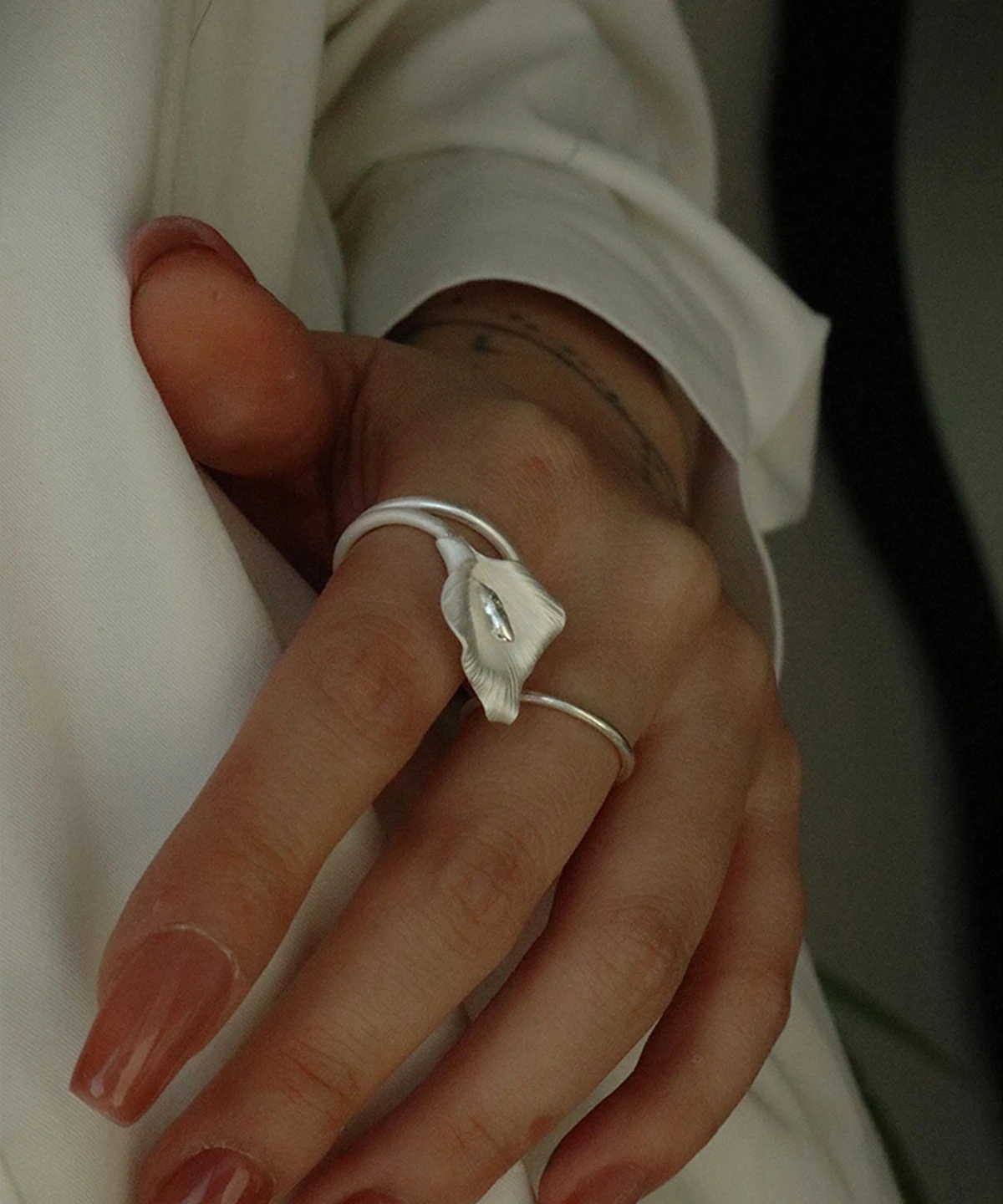 A close-up photograph showcases a hand wearing the Calla Lily Ring, a handmade piece crafted from 999 silver. The ring features an elegant design inspired by the calla lily flower, with a gracefully curved structure that resembles a blooming petal. The silver surface has a refined texture, enhancing its organic and artistic appearance. The adjustable band ensures a comfortable fit for various finger sizes. The hand is set against a soft white fabric background, complementing the delicate and sophisticated aesthetic of the jewelry. This image captures the timeless elegance of the Calla Lily Ring, making it a stunning accessory for jewelry lovers
