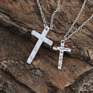 Two handcrafted 999 silver cross pendants rest upon a textured rock surface. The larger pendant features a dynamic flame-like pattern, while the smaller one showcases a hammered texture. Suspended from delicate silver chains, their contrasting finishes and intricate artistry stand out against the rugged stone backdrop, emphasizing their craftsmanship and elegance.
