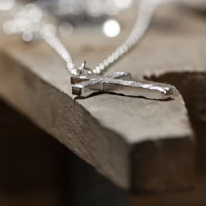 A close-up view of a handcrafted 999 silver cross pendant necklace resting on a wooden surface. The pendant features a textured, hammered finish that underscores its artisanal charm, suspended from a delicate silver chain. The softly blurred background draws attention to its intricate details and refined craftsmanship.