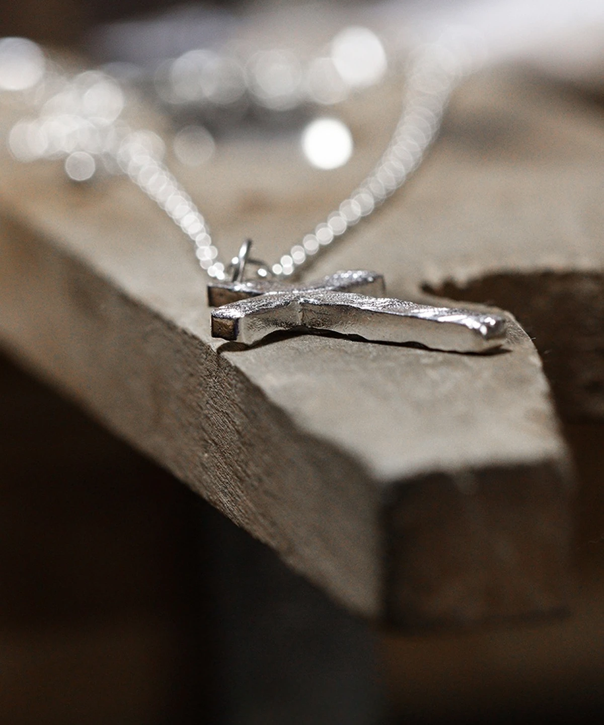 A close-up view of a handcrafted 999 silver cross pendant necklace resting on a wooden surface. The pendant features a textured, hammered finish that underscores its artisanal charm, suspended from a delicate silver chain. The softly blurred background draws attention to its intricate details and refined craftsmanship.