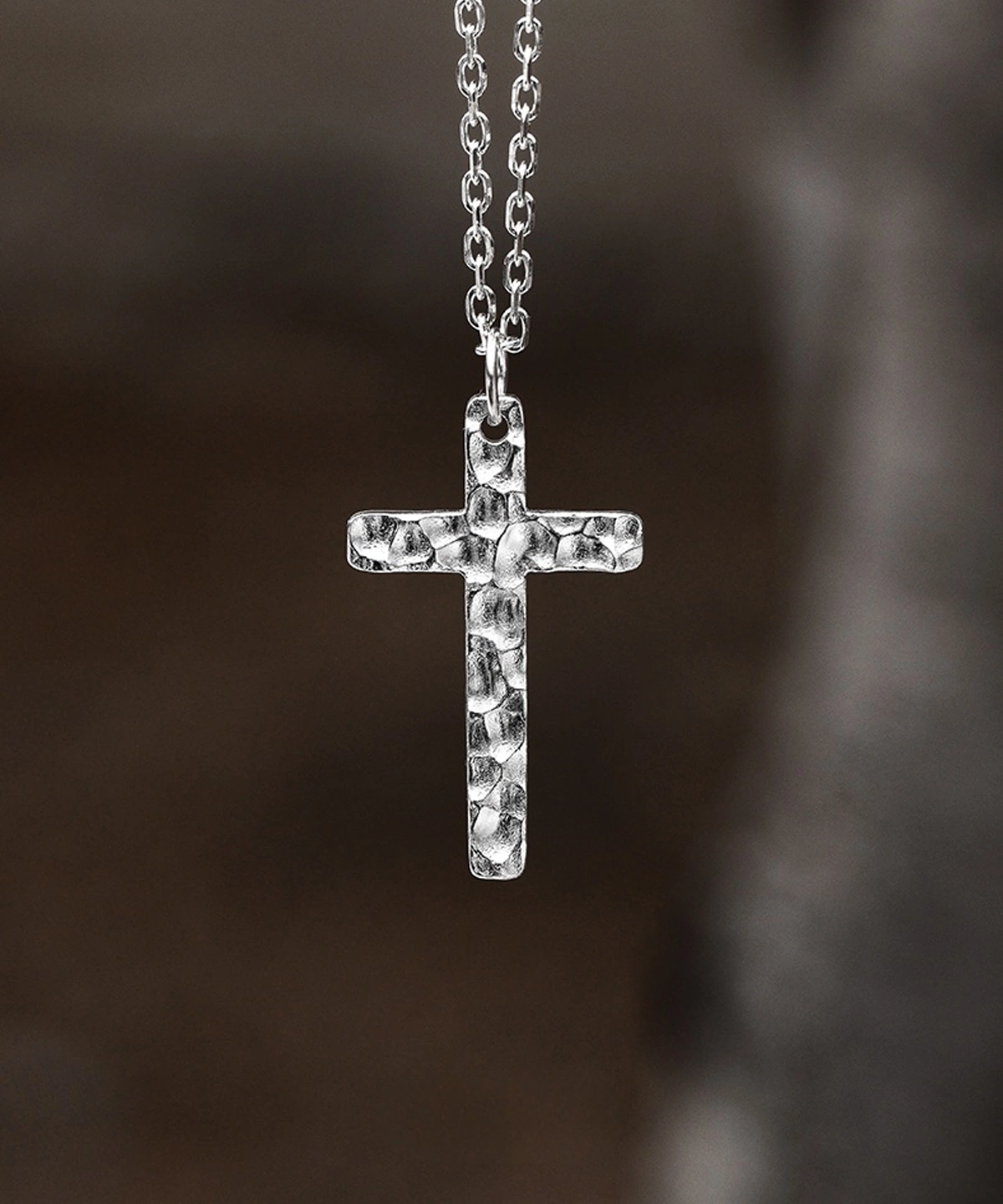 Handcrafted from 999 silver, this cross pendant features a textured, hammered finish that enhances its artisanal appeal. Suspended from an oval-link silver chain, the piece exudes understated elegance. The subtle imperfections in the metalwork highlight its handmade nature, making each pendant unique. The blurred background draws focus to the pendant’s intricate details, emphasizing its craftsmanship and refined simplicity.