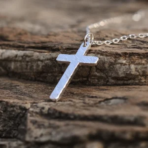 Handcrafted from 999 silver, this cross pendant features a textured surface that enhances its artisanal charm. Suspended from a delicate silver chain, it embodies elegance and craftsmanship. The rough stone background contrasts with the pendant’s polished finish, highlighting its intricate design. Ideal for those seeking a refined yet bold statement piece.