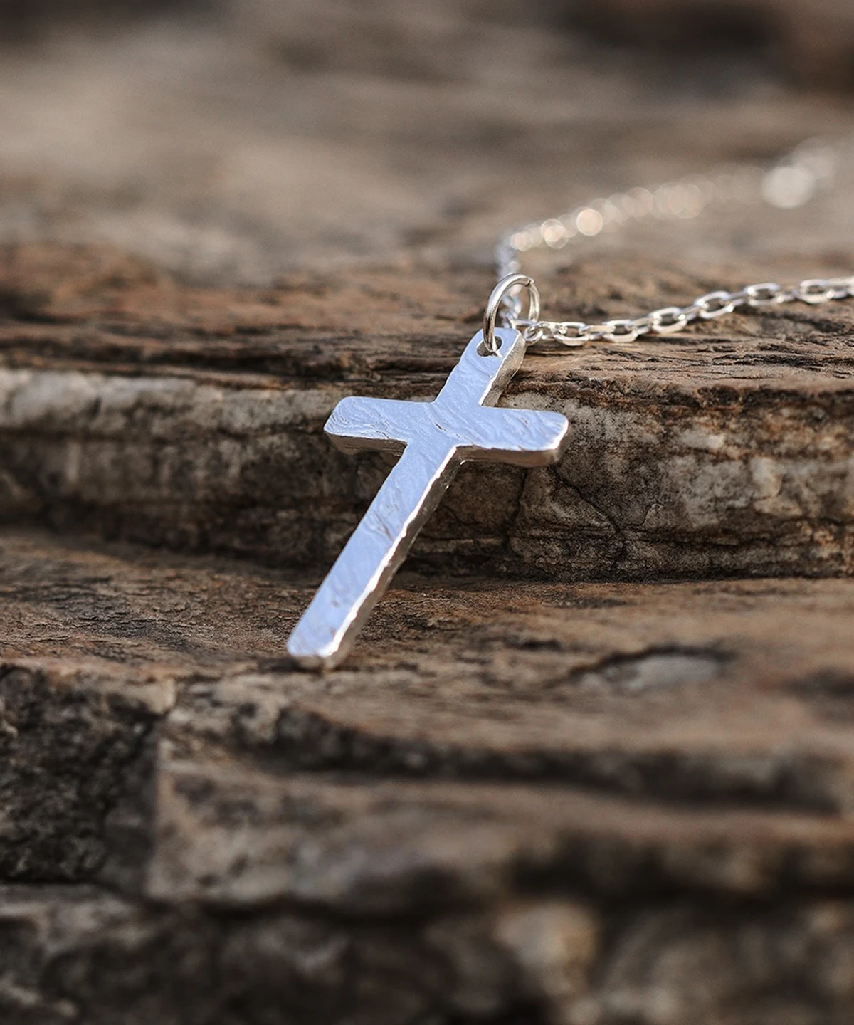 Handcrafted from 999 silver, this cross pendant features a textured surface that enhances its artisanal charm. Suspended from a delicate silver chain, it embodies elegance and craftsmanship. The rough stone background contrasts with the pendant’s polished finish, highlighting its intricate design. Ideal for those seeking a refined yet bold statement piece.