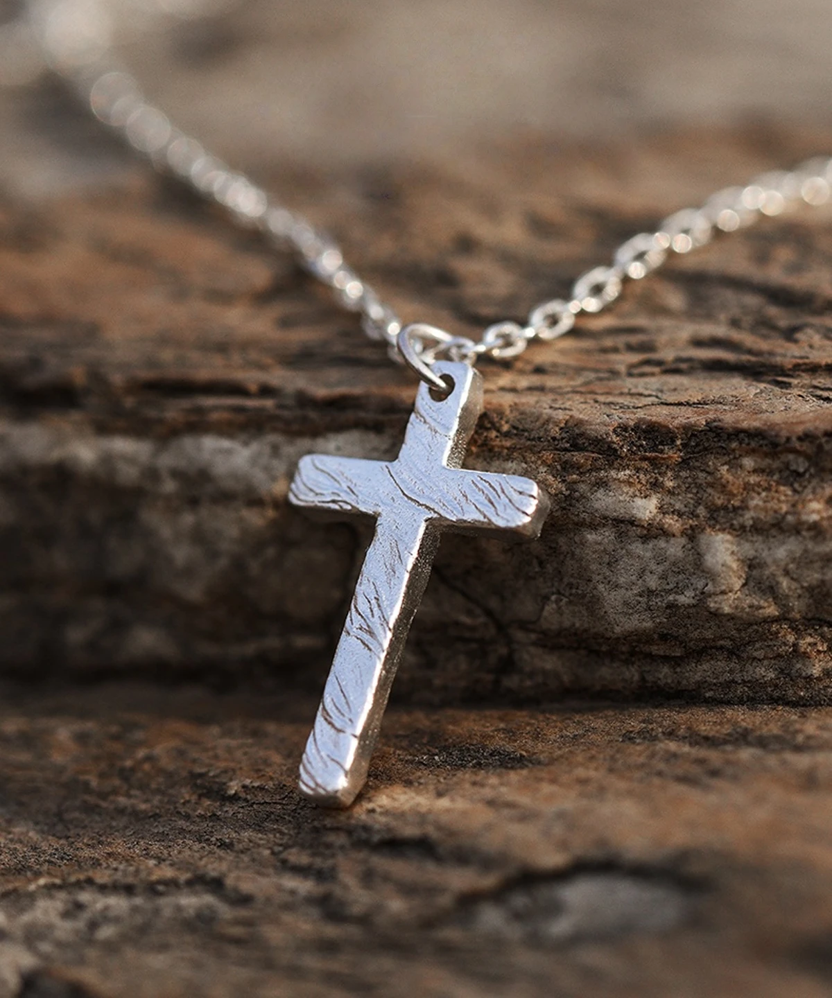 Handcrafted from 999 silver, this cross pendant features a textured surface that enhances its artisanal charm. Suspended from a delicate silver chain, it embodies elegance and craftsmanship. The rough stone background contrasts with the pendant’s polished finish, highlighting its intricate design. Ideal for those seeking a refined yet bold statement piece.