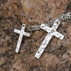 Two handcrafted 999 silver cross pendants rest upon a textured rock surface. The larger pendant features a dynamic flame-like pattern, while the smaller one showcases a hammered texture. Suspended from delicate silver chains, their contrasting finishes and intricate artistry stand out against the rugged stone backdrop, emphasizing their craftsmanship and elegance.
