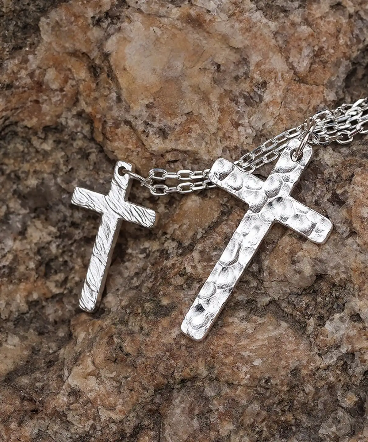 Two handcrafted 999 silver cross pendants rest upon a textured rock surface. The larger pendant features a dynamic flame-like pattern, while the smaller one showcases a hammered texture. Suspended from delicate silver chains, their contrasting finishes and intricate artistry stand out against the rugged stone backdrop, emphasizing their craftsmanship and elegance.