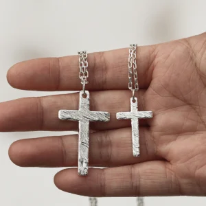 A close-up shot captures a hand elegantly holding two handcrafted silver cross necklaces. The larger pendant rests between the thumb and index finger, while the smaller one is nestled between the index and middle fingers. Both crosses display intricate textured details and are connected by delicate silver chains, all set against a soft, light backdrop that accentuates their refined craftsmanship.