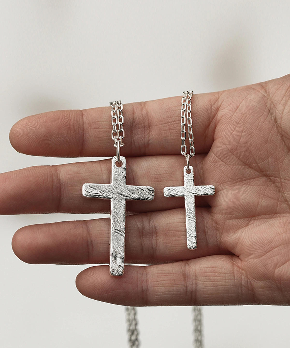A close-up shot captures a hand elegantly holding two handcrafted silver cross necklaces. The larger pendant rests between the thumb and index finger, while the smaller one is nestled between the index and middle fingers. Both crosses display intricate textured details and are connected by delicate silver chains, all set against a soft, light backdrop that accentuates their refined craftsmanship.