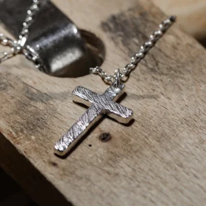Handcrafted from 999 silver, this cross pendant boasts a textured surface that enhances its artisanal appeal. Suspended from a sleek silver chain, it exudes timeless elegance and craftsmanship. The rustic wooden background adds depth to the piece, emphasizing its handmade nature. Perfect for those who appreciate high-quality, unique jewelry with a refined yet bold aesthetic.