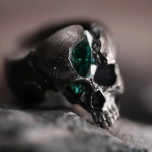 A close-up shot of the Dark Skull Ring in S925 silver, featuring a bold skull motif with a striking green gemstone inlay. The ring’s dark, oxidized finish enhances its edgy, gothic appeal, and it is displayed on a textured surface that complements its rugged aesthetic.