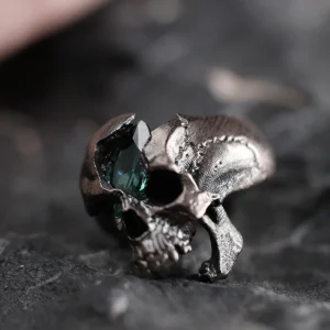 A close-up shot of the Dark Skull Ring in S925 silver, featuring a bold skull motif with a striking green gemstone inlay. The ring’s dark, oxidized finish enhances its edgy, gothic appeal, and it is displayed on a textured surface that complements its rugged aesthetic.