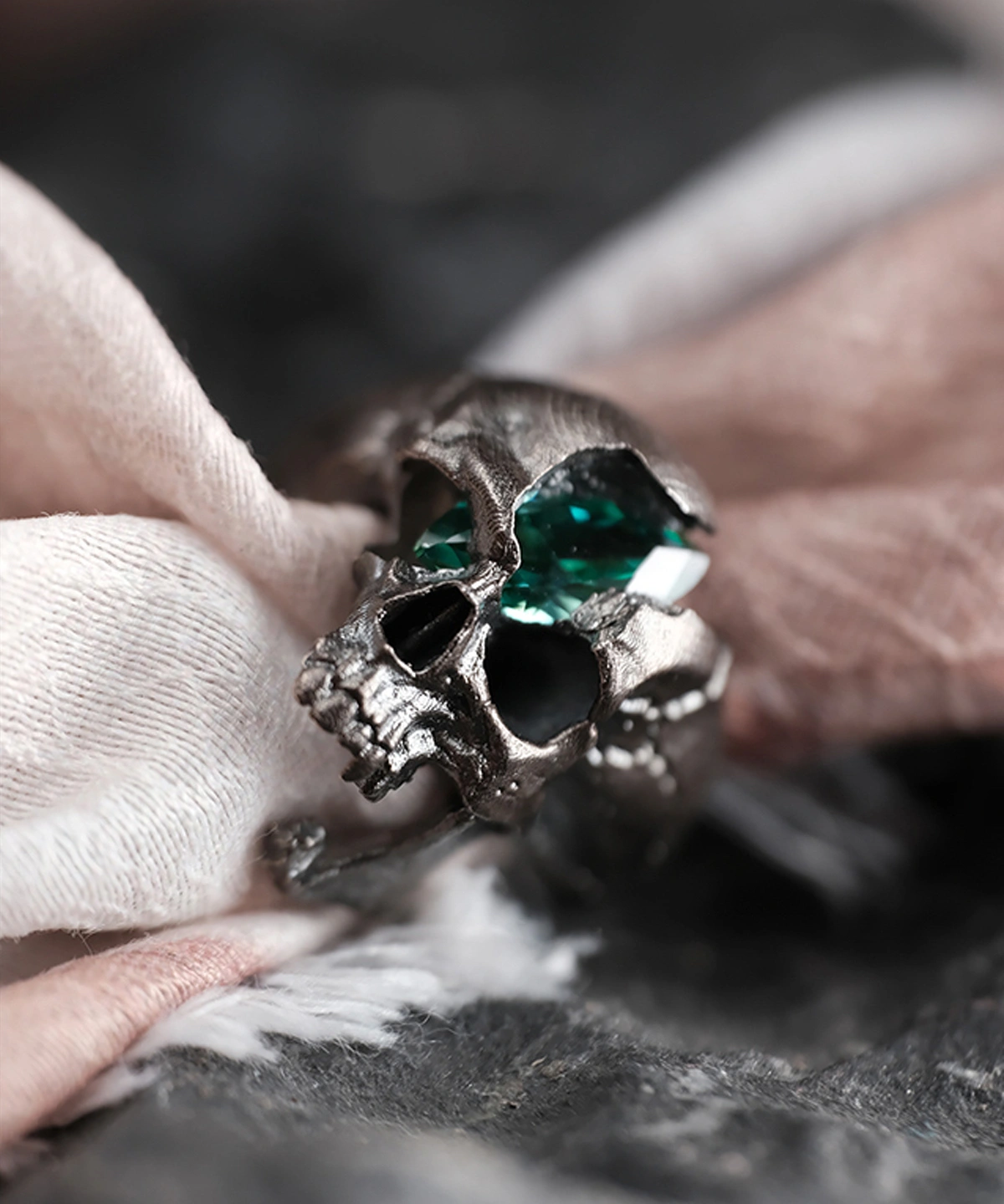 A close-up image of the Dark Skull Ring, crafted from S925 silver. The ring showcases a bold skull design with intricate detailing, featuring a striking green gemstone inlay set in the eye sockets. The polished silver and dark oxidized finish create a dramatic contrast, emphasizing the edgy, gothic aesthetic.