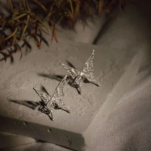 A pair of butterfly earrings and ear cuffs, designed without the need for piercings, elegantly rest on a textured concrete block. The earrings feature intricately crafted silver butterflies, their delicate wings casting soft shadows. Surrounding the display are slender branches with leaves, adding a touch of nature-inspired sophistication.