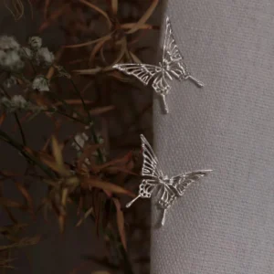 The Dream Butterfly Earrings & Ear Cuffs are elegantly displayed on a soft-textured fabric, their delicate silver butterflies capturing light with refined detail. Designed for effortless wear without piercings, these butterfly earrings feature graceful wing patterns that evoke a sense of ethereal beauty. Dried flowers surround the jewelry, adding an organic charm to the composition.