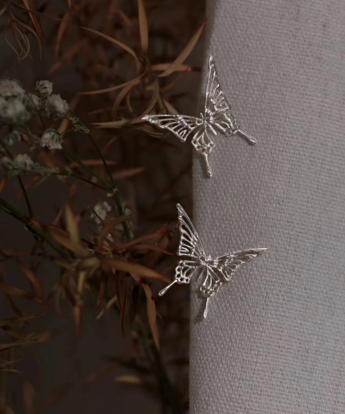 The Dream Butterfly Earrings & Ear Cuffs are elegantly displayed on a soft-textured fabric, their delicate silver butterflies capturing light with refined detail. Designed for effortless wear without piercings, these butterfly earrings feature graceful wing patterns that evoke a sense of ethereal beauty. Dried flowers surround the jewelry, adding an organic charm to the composition.