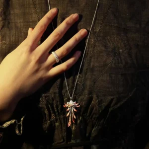 Close-up image of a model’s hand resting on dark textured fabric, highlighting the Drooping Blossom 925 Sterling Silver Long Necklace. The necklace features a delicate pendant resembling a stylized leaf with intricate details in mixed silver and copper tones, draped elegantly from a thin silver chain. A silver ring on the middle finger adds a modern touch, emphasizing the piece’s refined craftsmanship. Ideal for those seeking sophisticated, fine jewelry for both casual and formal occasions.
