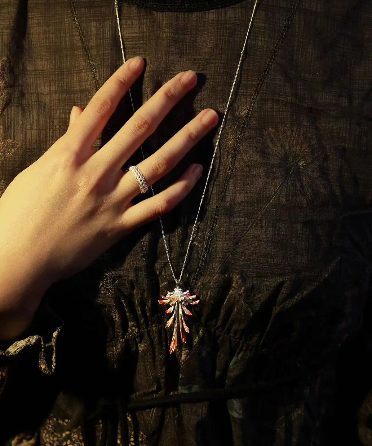 Close-up image of a model’s hand resting on dark textured fabric, highlighting the Drooping Blossom 925 Sterling Silver Long Necklace. The necklace features a delicate pendant resembling a stylized leaf with intricate details in mixed silver and copper tones, draped elegantly from a thin silver chain. A silver ring on the middle finger adds a modern touch, emphasizing the piece’s refined craftsmanship. Ideal for those seeking sophisticated, fine jewelry for both casual and formal occasions.