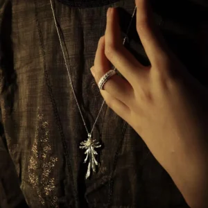 Drooping Blossom | 925 Sterling Silver Long Necklace features a refined, drooping floral pendant crafted in sterling silver. The necklace is displayed on a model wearing a dark, textured garment with intricate patterns under dramatic, moody lighting. A subtle silver ring on the model's index finger softly touches the pendant, adding a modern accent. This elegant piece is perfect for both formal events and daily wear, combining timeless sophistication with contemporary design.