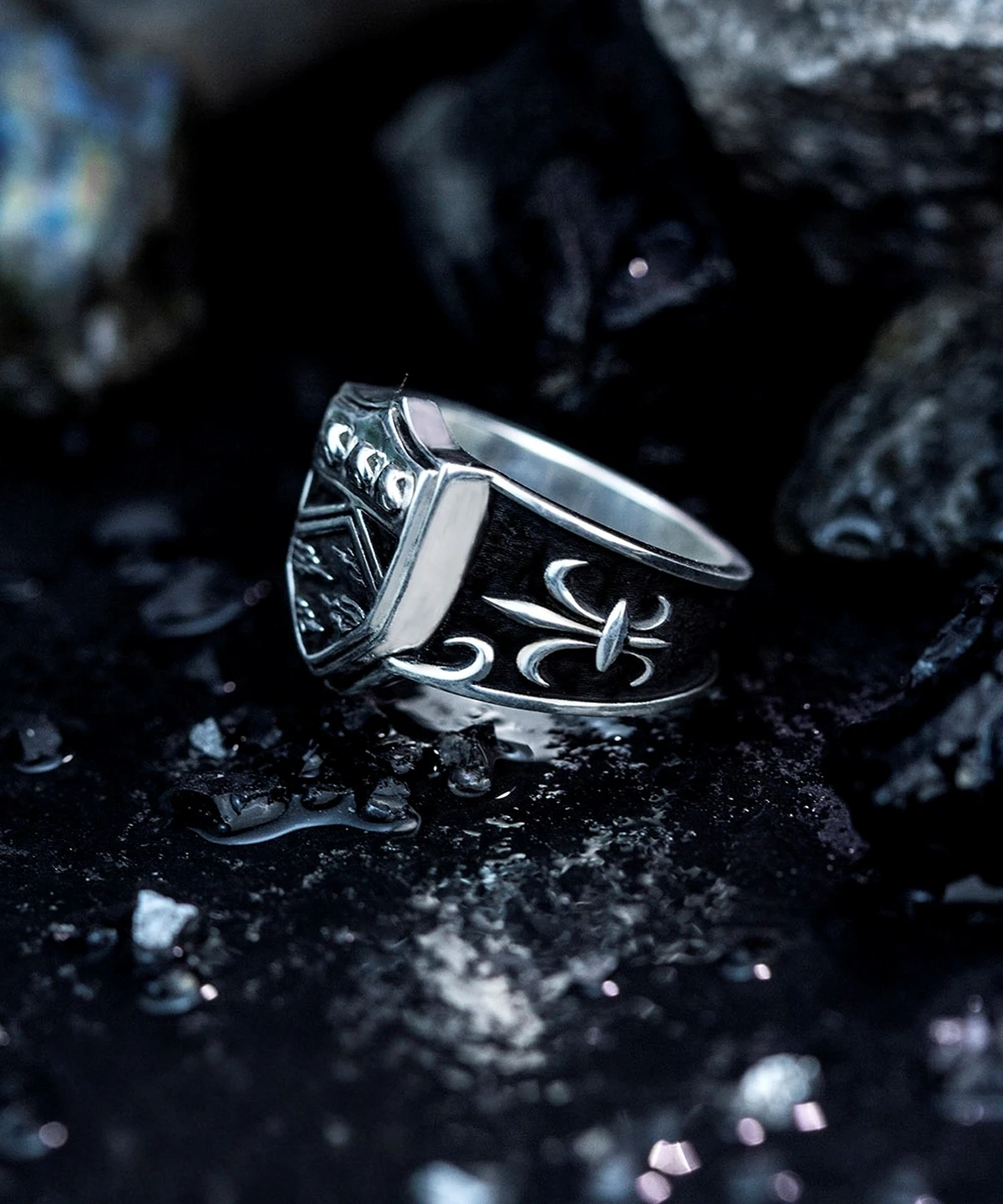 A close-up view of the Katekyo Hitman Reborn Vongola Ring, crafted in silver with intricate engravings, presented against a dark, textured background with scattered reflective particles.