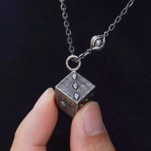 Close-up view of the Prison Realm Dice Necklace—a handcrafted sterling silver piece. The image shows a hand holding a cube pendant, intricately decorated with diamond-like stones on its visible faces. A unique chain with an eye-shaped detail completes the design, set against a dark backdrop that accentuates the textured finish and artisanal craftsmanship.
