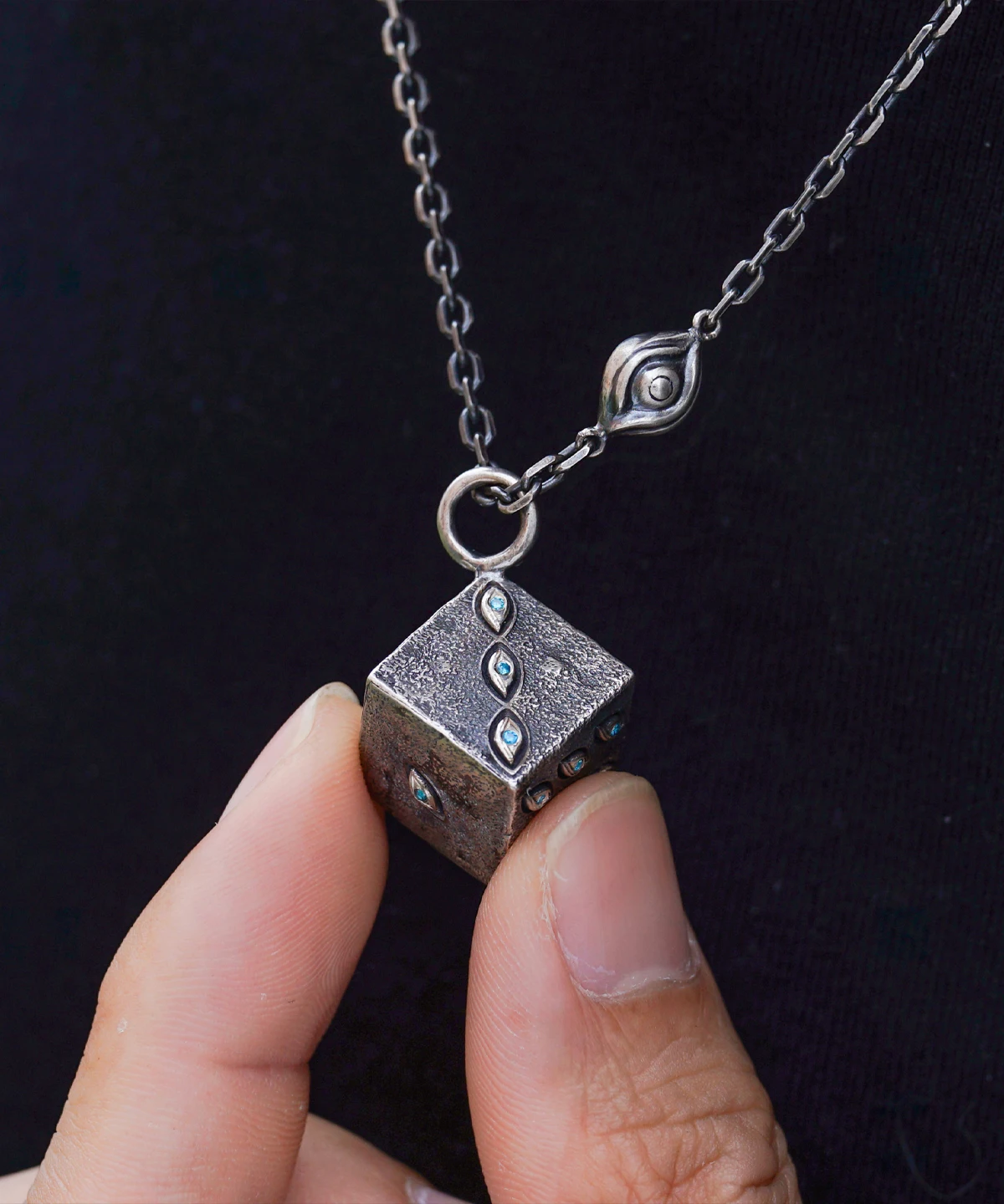 Close-up view of the Prison Realm Dice Necklace—a handcrafted sterling silver piece. The image shows a hand holding a cube pendant, intricately decorated with diamond-like stones on its visible faces. A unique chain with an eye-shaped detail completes the design, set against a dark backdrop that accentuates the textured finish and artisanal craftsmanship.