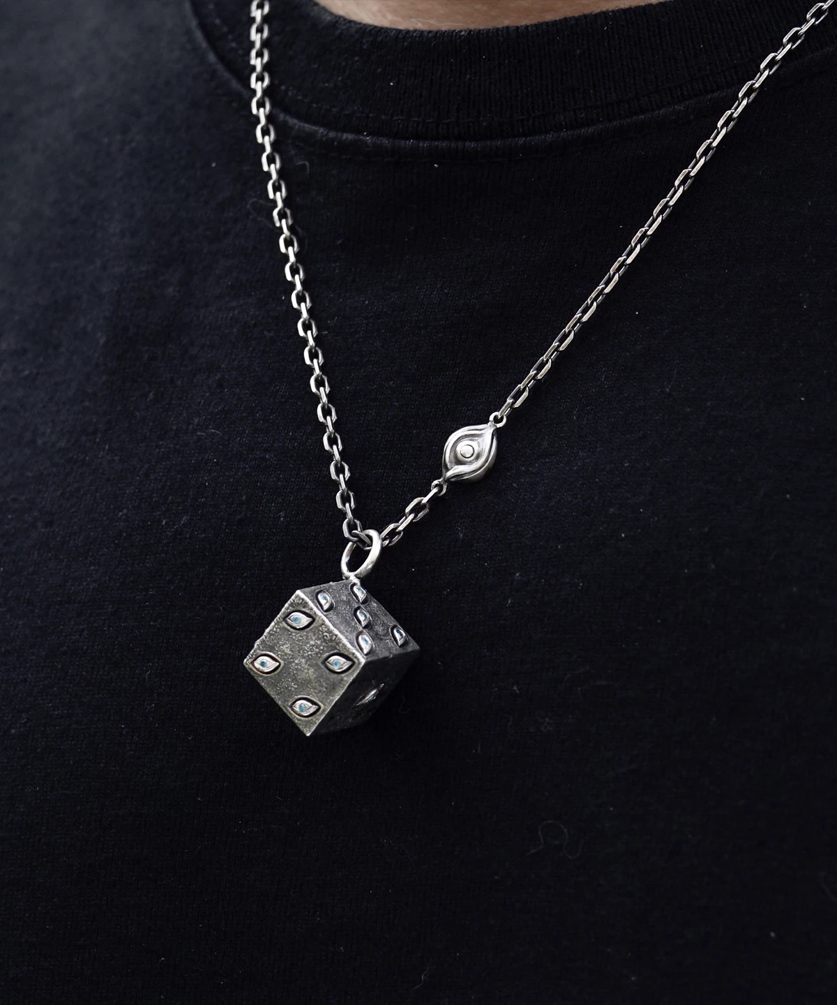 This handcrafted sterling silver necklace features a dice-inspired cube pendant with a rough texture and subtle eye-shaped engravings. The twisted metallic chain, enhanced by a delicate eye motif, complements the pendant's artistic design. Set against a dark backdrop, the piece highlights intricate craftsmanship and a unique, symbolic aesthetic.