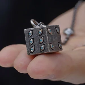 A close-up view of the Prison Realm Dice Necklace—a handcrafted sterling silver piece. A hand holds the chain featuring a six-sided metallic die pendant, uniquely adorned with eye-shaped engravings on each face. The dark background enhances the intricate details and reflective texture of the pendant and chain, emphasizing the artisanal quality of this distinctive design.