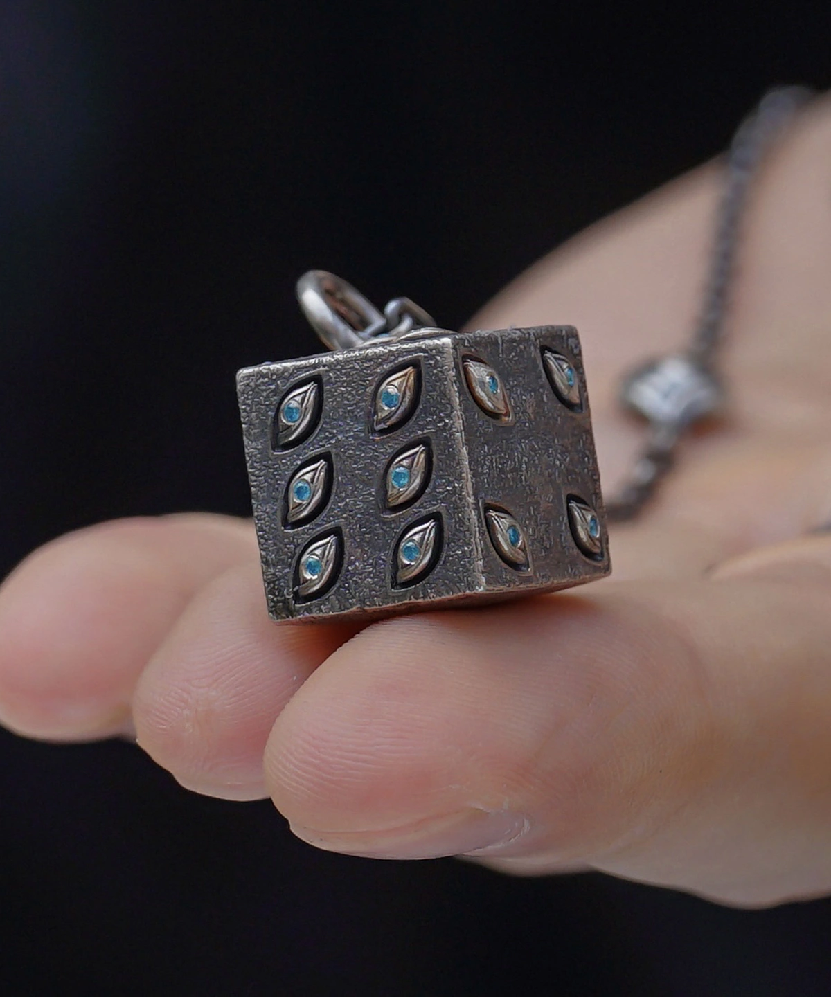 A close-up view of the Prison Realm Dice Necklace—a handcrafted sterling silver piece. A hand holds the chain featuring a six-sided metallic die pendant, uniquely adorned with eye-shaped engravings on each face. The dark background enhances the intricate details and reflective texture of the pendant and chain, emphasizing the artisanal quality of this distinctive design.