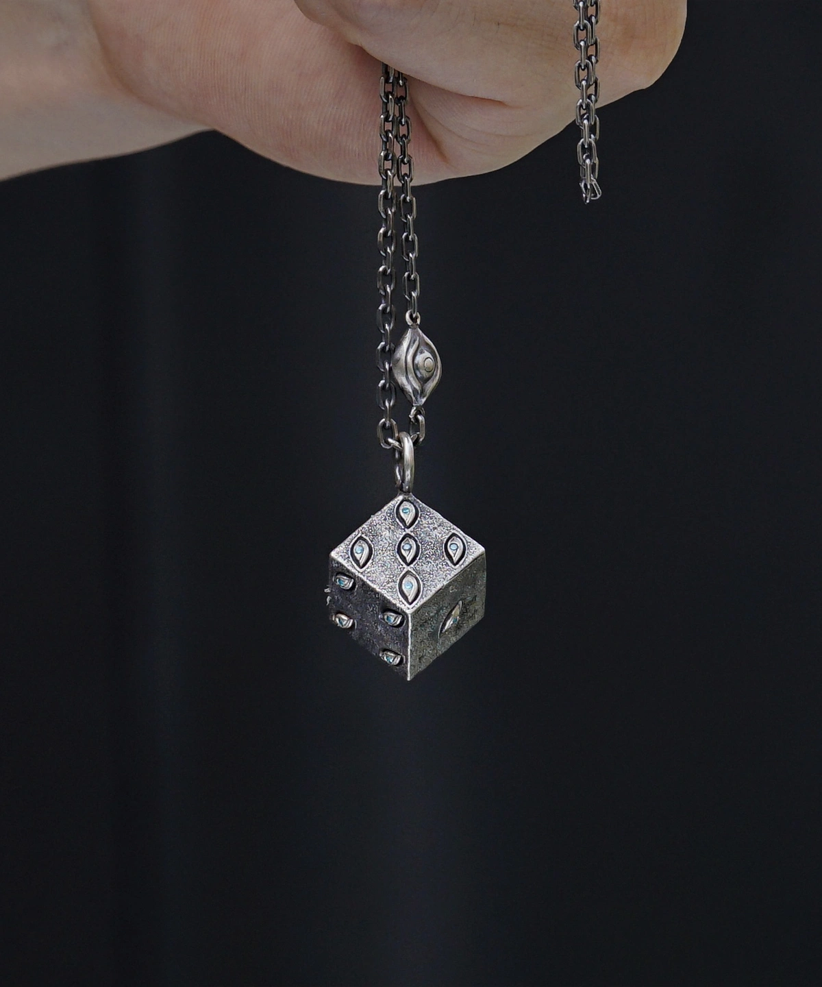 A close-up view of a handcrafted sterling silver necklace featuring a unique six-sided dice pendant. The metallic dice is elegantly adorned with intricate engravings and gem-like accents that add a refined touch. A medium-thickness chain complements the pendant, and the dark background highlights its lustrous texture. This Prison Realm Dice Necklace showcases a striking blend of bold design and skilled, artisanal craftsmanship.