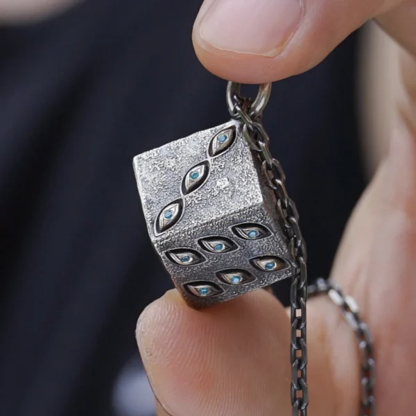 The image displays a close-up of a hand holding a handcrafted sterling silver necklace featuring a cube-shaped pendant. The pendant’s textured surface is adorned with subtle, eye-shaped engravings and small blue stones. A delicate chain is visible, perfectly complementing the unique design of the Prison Realm Dice Necklace. This view emphasizes the exquisite craftsmanship and artistic detail of the piece.
