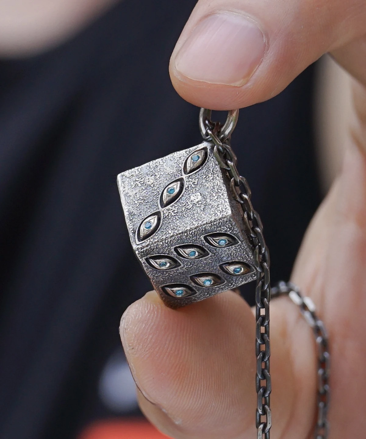 The image displays a close-up of a hand holding a handcrafted sterling silver necklace featuring a cube-shaped pendant. The pendant’s textured surface is adorned with subtle, eye-shaped engravings and small blue stones. A delicate chain is visible, perfectly complementing the unique design of the Prison Realm Dice Necklace. This view emphasizes the exquisite craftsmanship and artistic detail of the piece.
