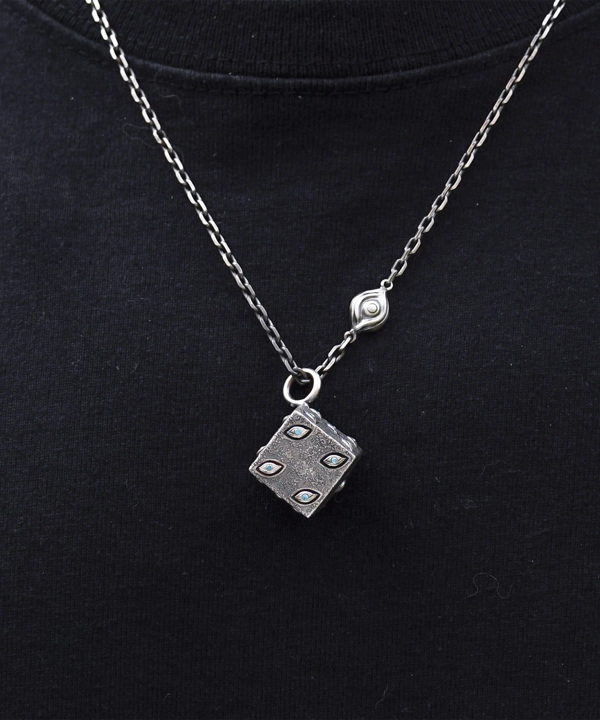 This handcrafted sterling silver necklace features a dice-inspired cube pendant with a rough texture and subtle eye-shaped engravings. The twisted metallic chain, enhanced by a delicate eye motif, complements the pendant's artistic design. Set against a dark backdrop, the piece highlights intricate craftsmanship and a unique, symbolic aesthetic.