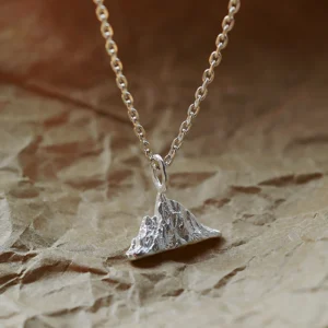 A close-up image of the Snow Mountain Necklace, featuring a 999 pure silver pendant shaped like a majestic mountain range. The intricate peaks and valleys of the pendant evoke the beauty of snow-covered summits, suspended from a delicate silver chain. The background is a textured, warm-toned surface that enhances the brilliance of the piece.