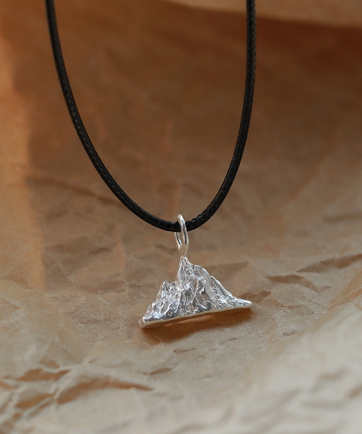 A close-up of the Snow Mountain Necklace featuring a 999 pure silver pendant intricately shaped like a mountain. The pendant hangs from a black cord and is displayed on a textured brown background, enhancing its rustic elegance.