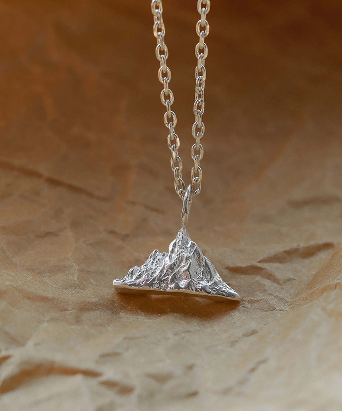 A close-up image of the Snow Mountain Necklace, featuring a 999 pure silver pendant shaped like a majestic mountain range. The intricate peaks and valleys of the pendant evoke the beauty of snow-covered summits, suspended from a delicate silver chain. The background is a textured, warm-toned surface that enhances the brilliance of the piece.