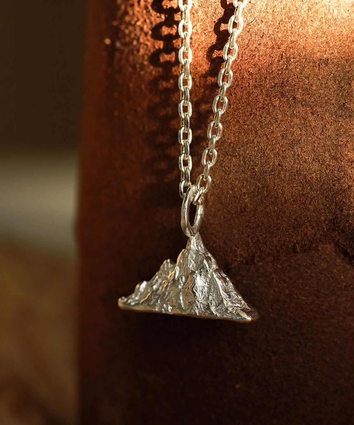 A close-up image of a Snow Mountain Necklace featuring a 999 pure silver pendant. The pendant is shaped like a mountain range with detailed textures, hanging from a silver chain against a brown background.