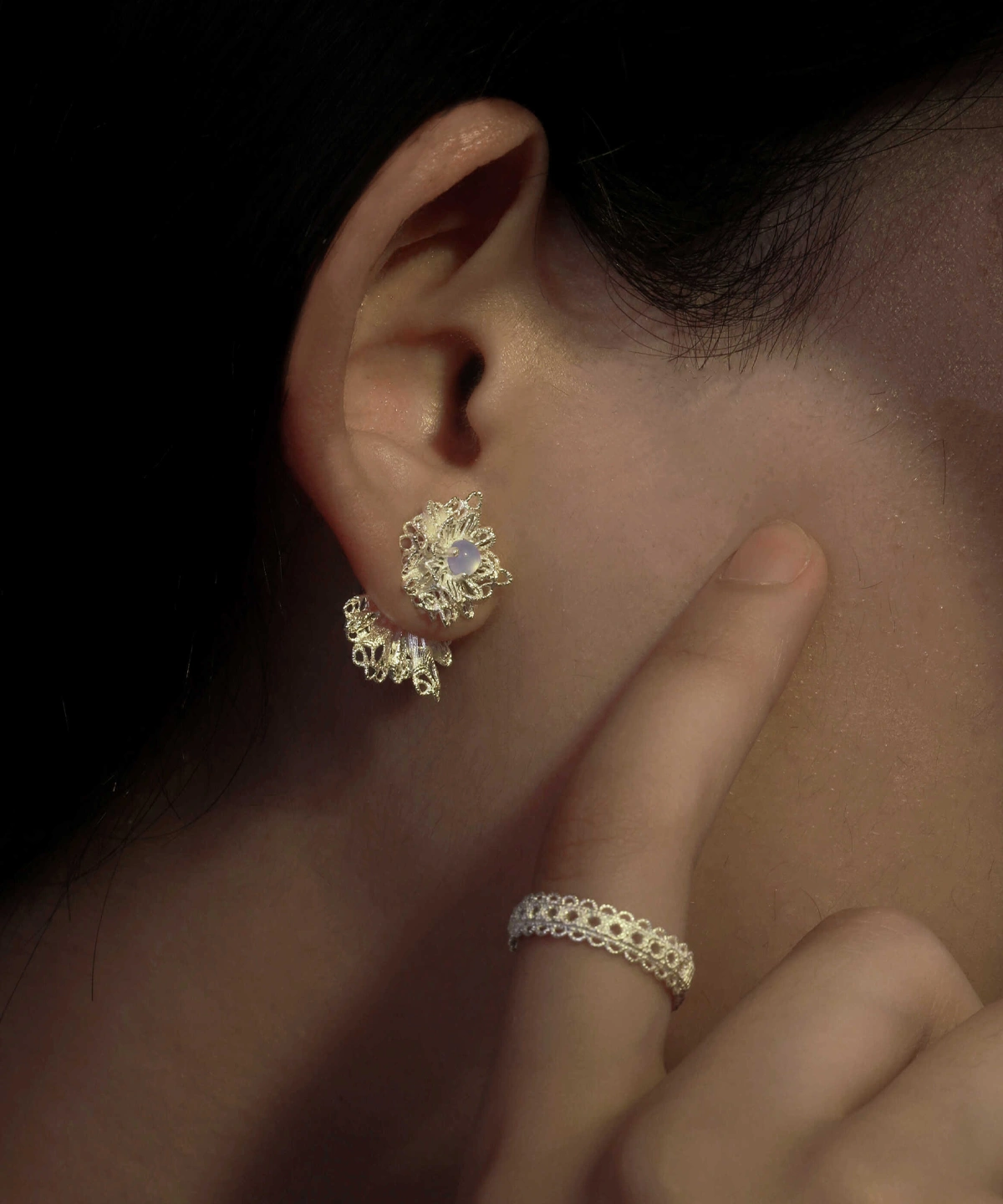A close-up of an ear adorned with Snowflake Earrings. The earrings are 925 Sterling Silver Lace Stud Earrings, featuring an intricate lace-like snowflake design with a central gemstone. A delicate finger points toward the earrings, complementing the look with a matching lace-patterned ring.