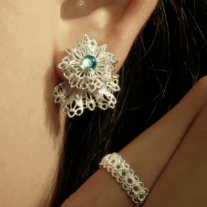 A close-up of a person's ear adorned with Snowflake Earrings made of 925 Sterling Silver Lace. The earrings feature intricate snowflake-inspired lacework with a central blue gemstone. A matching lace-patterned ring is also visible near the ear, complementing the elegant winter motif.