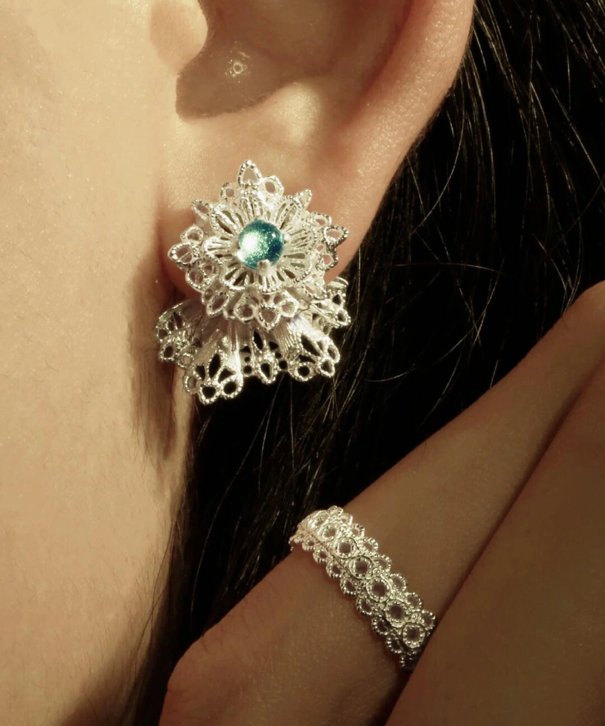 A close-up of a person's ear adorned with Snowflake Earrings made of 925 Sterling Silver Lace. The earrings feature intricate snowflake-inspired lacework with a central blue gemstone. A matching lace-patterned ring is also visible near the ear, complementing the elegant winter motif.