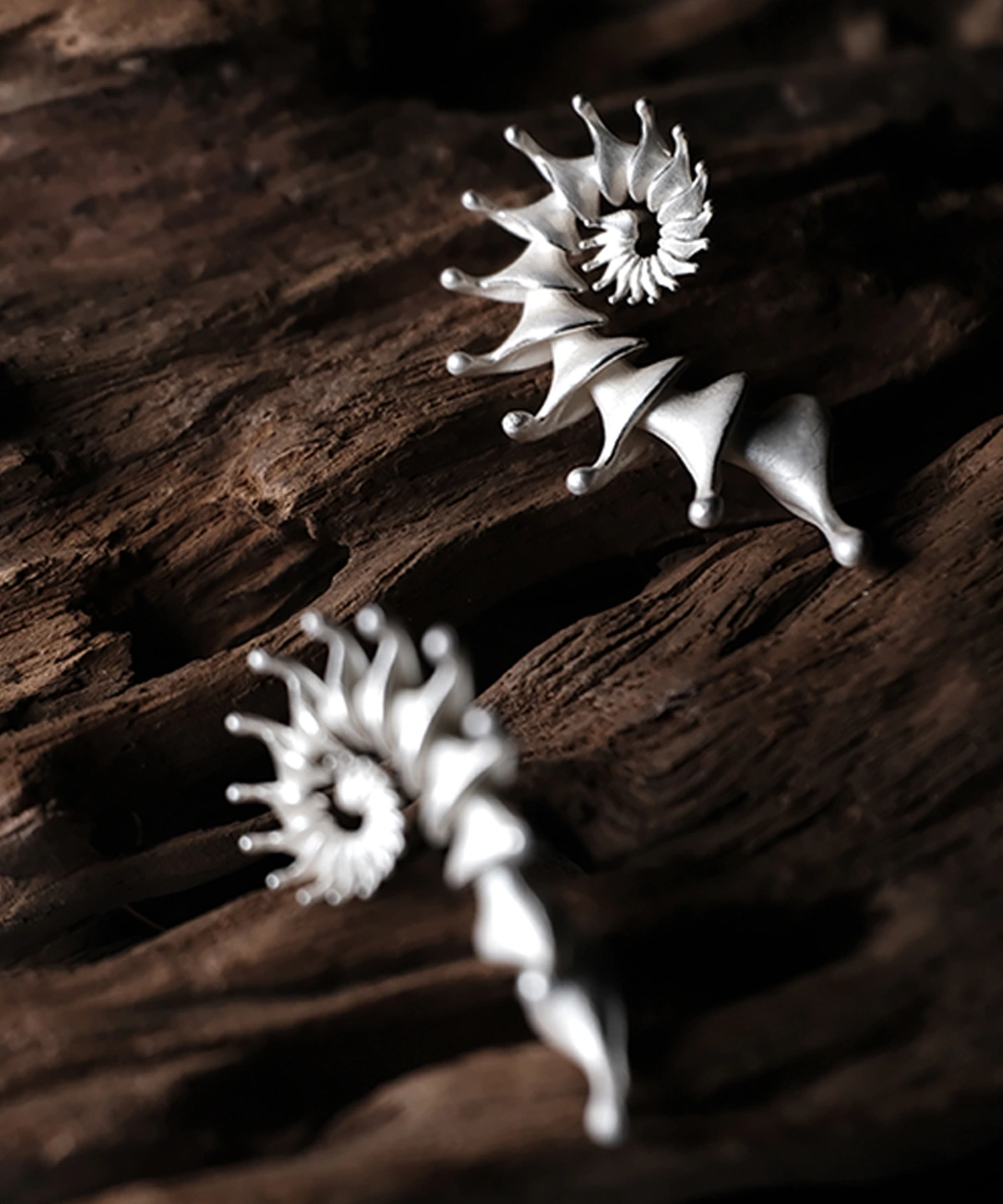 A close-up image of two Spiral Earrings made from S925 sterling silver, designed as Designer Ear Cuffs. The earrings feature an elegant, flowing spiral structure with smooth curves and pointed tips, presented against a rustic wooden background.