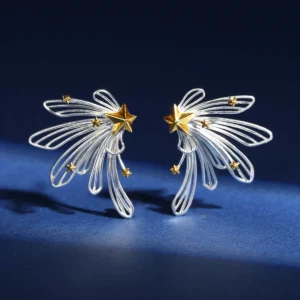 A close-up view shows sterling silver shooting star stud earrings from the Star Trail collection. Intricate wire loops form dynamic shooting star tails, each featuring a bold central gold star with scattered smaller gold accents. Set against a dark blue backdrop, the earrings exude a celestial, modern elegance.
