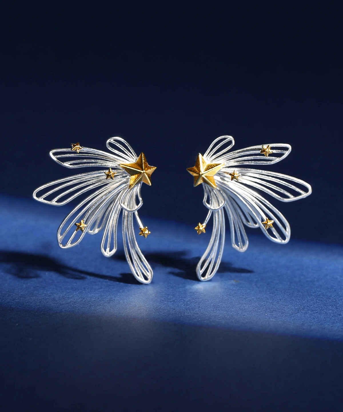 A close-up view shows sterling silver shooting star stud earrings from the Star Trail collection. Intricate wire loops form dynamic shooting star tails, each featuring a bold central gold star with scattered smaller gold accents. Set against a dark blue backdrop, the earrings exude a celestial, modern elegance.