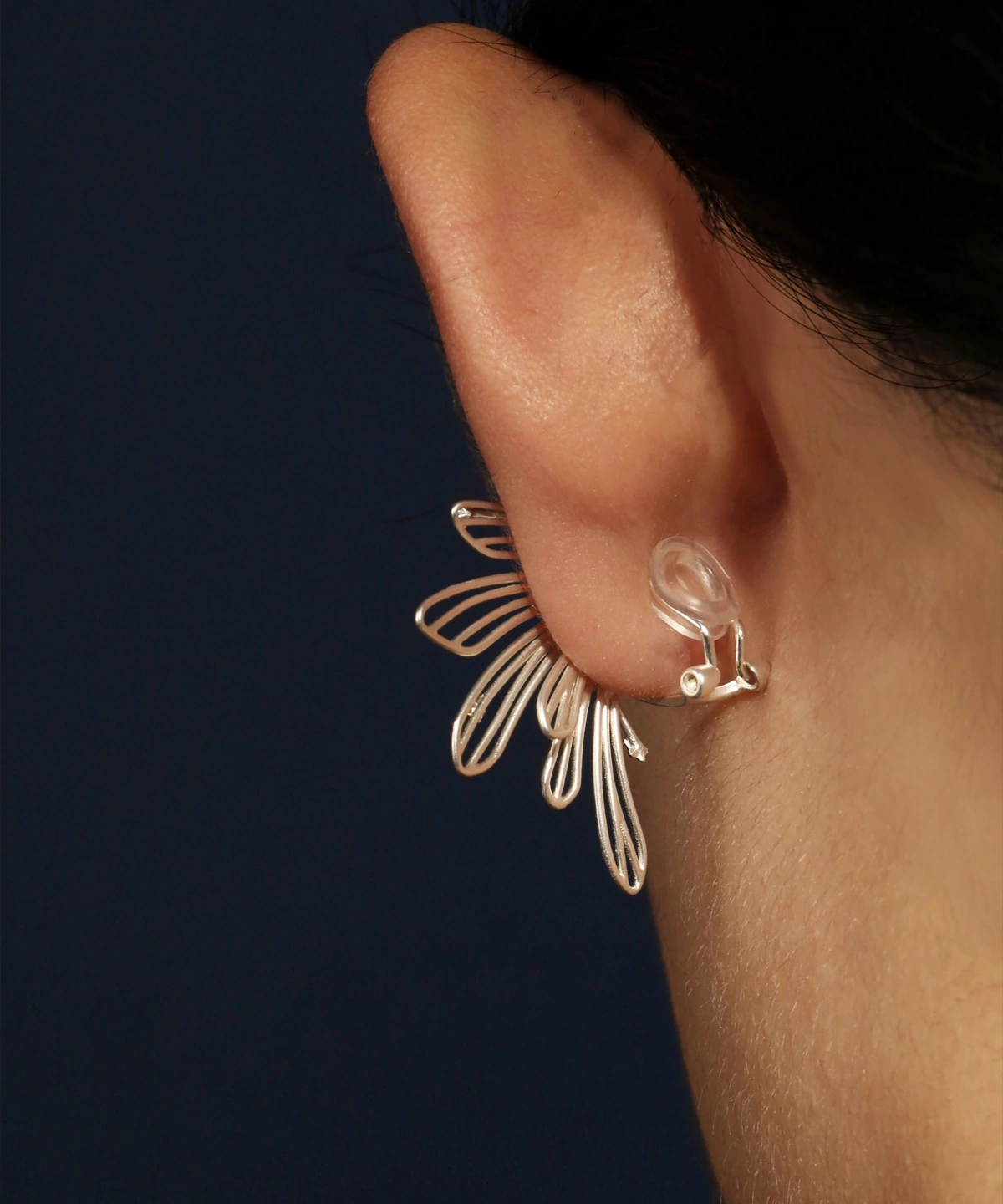 A close-up of the back of an ear showcases the Star Trail ear cuff, crafted from 925 sterling silver. The delicate design features a trail of small star motifs that elegantly wrap around the ear, creating a celestial-inspired look. The cuff is secured with a transparent clip, making it suitable for non-pierced ears. Its fine details and sleek silver finish enhance its sophisticated yet playful charm.